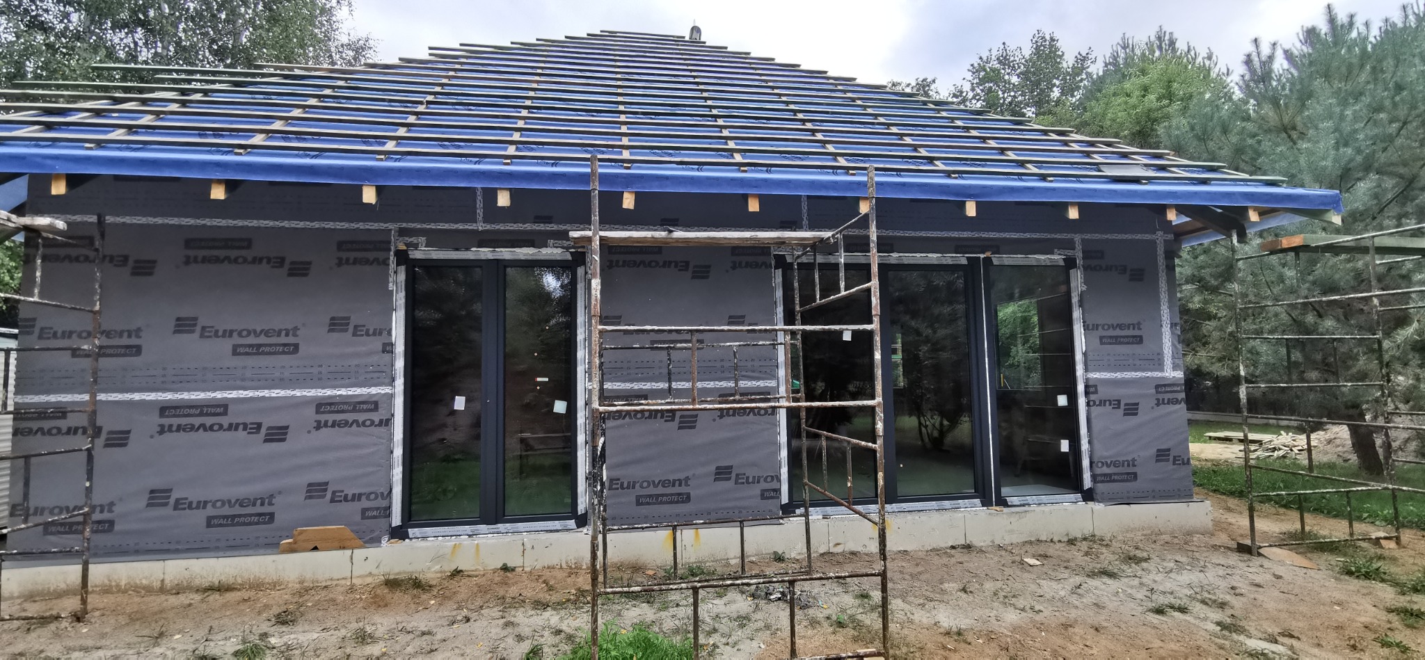 Budowa domu szkieletowego: widok na ściany z oknami i rusztowaniem, z membraną Eurovent. W tle drzewa i niebo. Etap montażu okien i izolacji.