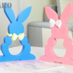 Dwa dekoracyjne, wycięte z drewna zajączki: niebieski z żółtą kokardką i różowy z białą kokardką na uszach, stojące na stole. W tle widoczny fragment sofy.