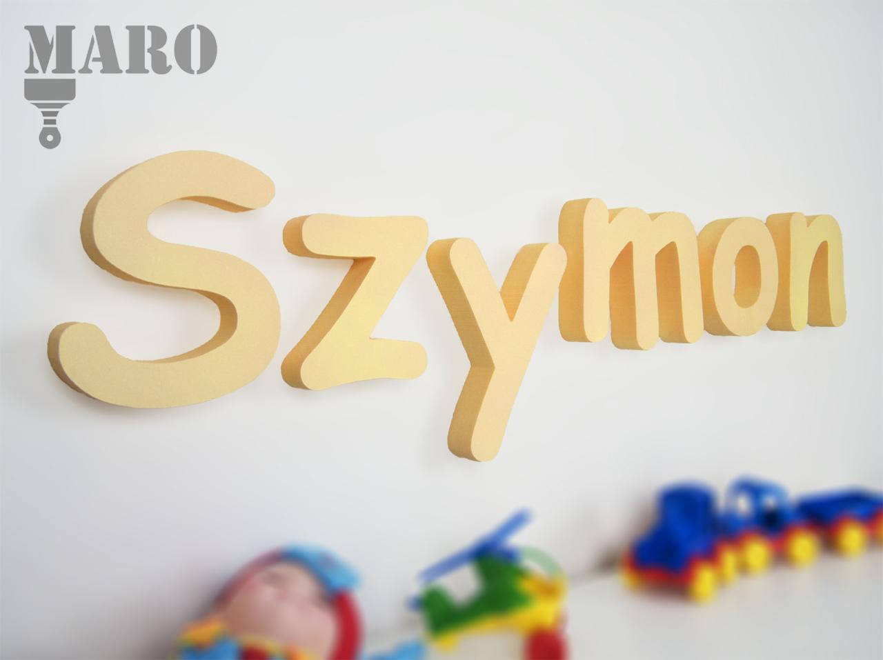 Imię Szymon, wykonane z jasnych, trójwymiarowych liter, umieszczone na białej ścianie, z rozmytymi zabawkami w tle i logo firmy w lewym górnym rogu.
