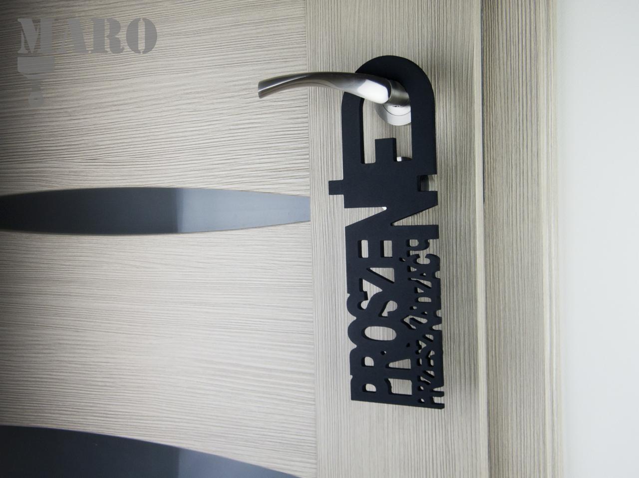 Czarny, wycinany laserowo zawieszka na klamkę drzwi z napisem 'Proszę nie przeszkadzać', wisząca na drzwiach z drewnopodobnym wzorem.