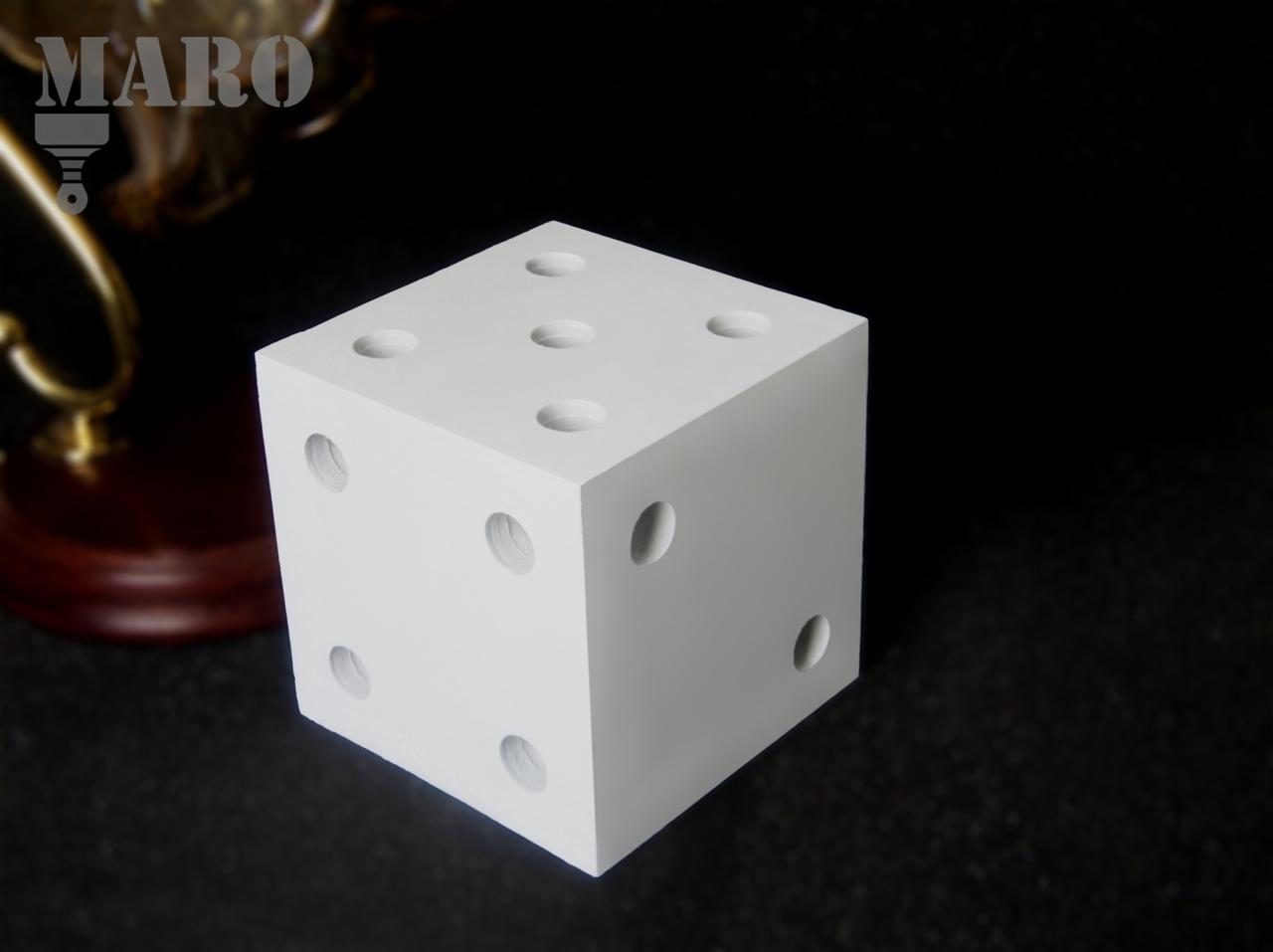 Biała, sześcienna kostka do gry z wytłoczonymi oczkami na ciemnym tle, obok dekoracyjna lampka z logo MARO.