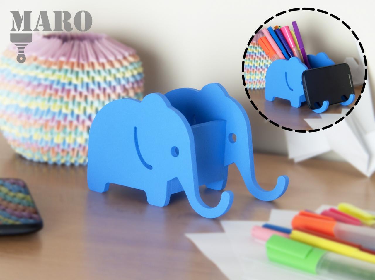 Niebieski organizer biurkowy w kształcie słonia, z przegródkami na długopisy i telefon, umieszczony na drewnianym biurku obok kolorowej lampki origami i zestawu markerów.