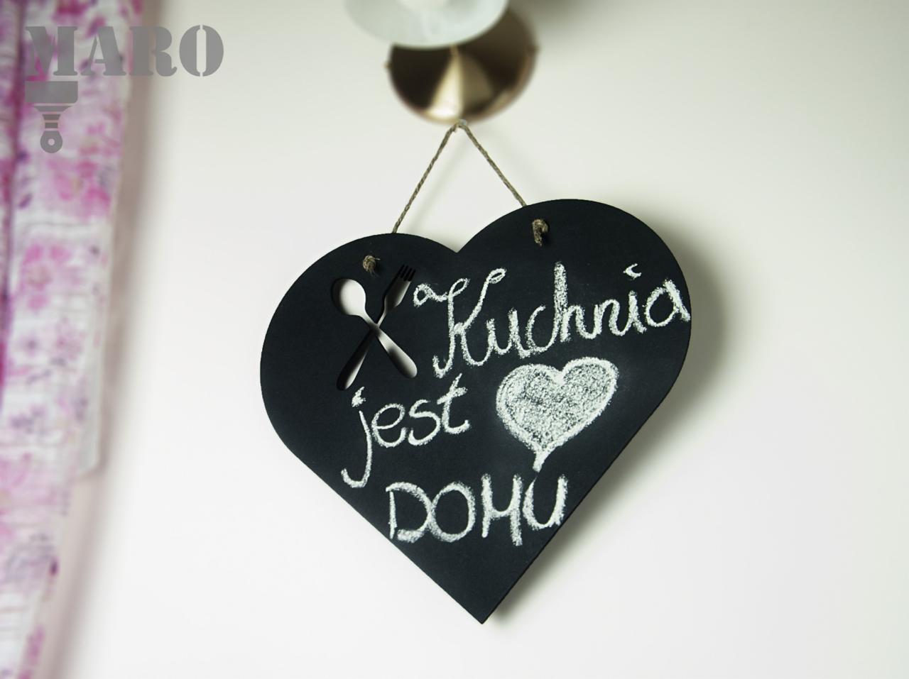 Tablica w kształcie serca z napisem 'Kuchnia jest DOMU', z wyciętym symbolem sztućców, zawieszona na sznurku, w tle fragment zasłony.