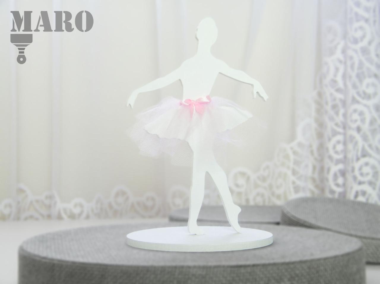 Biała, drewniana figurka baletnicy w tiulowej spódniczce z różową kokardką, ustawiona na szarym podeście, na tle białej, koronkowej zasłony.
