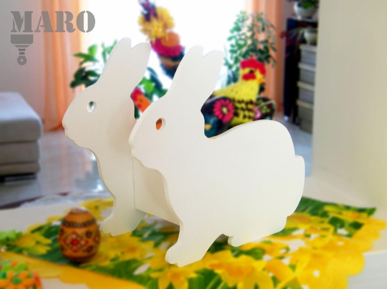 Dwa białe, drewniane zajączki wielkanocne na stole z żółtą serwetą w kwiaty, w tle pisanka i dekoracja z kurczakiem.