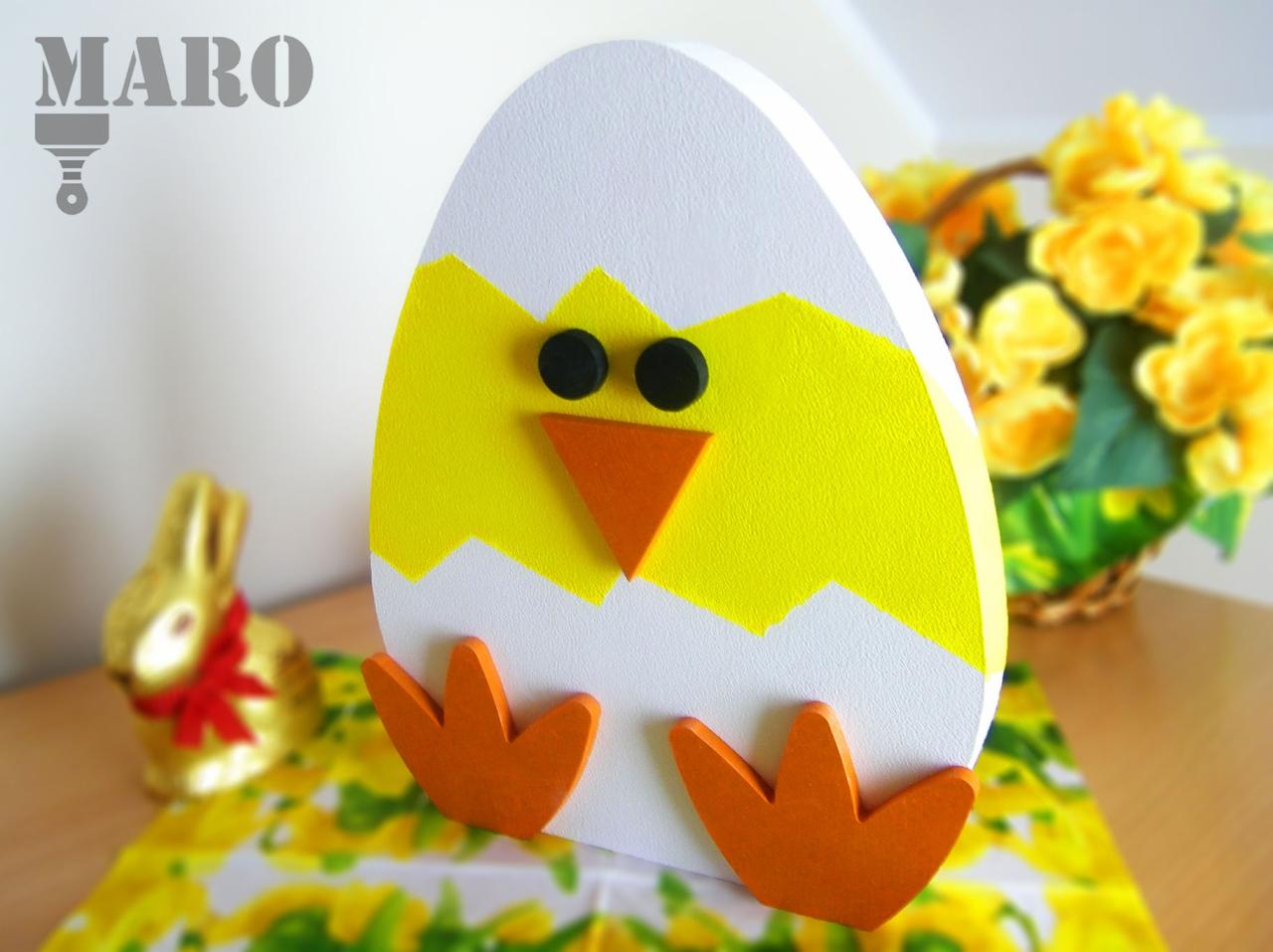 Ręcznie robiona drewniana ozdoba wielkanocna w kształcie pisklęcia wyłaniającego się z jajka, na stole z żółtymi kwiatami w tle i czekoladowym zającem.