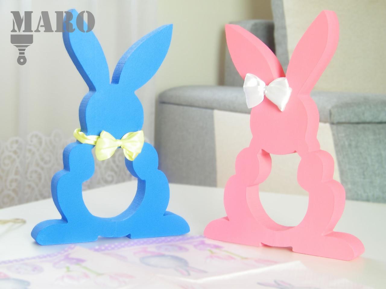 Dwa dekoracyjne, wycięte z drewna zajączki: niebieski z żółtą kokardką i różowy z białą kokardką na uszach, stojące na stole. W tle widoczny fragment sofy.