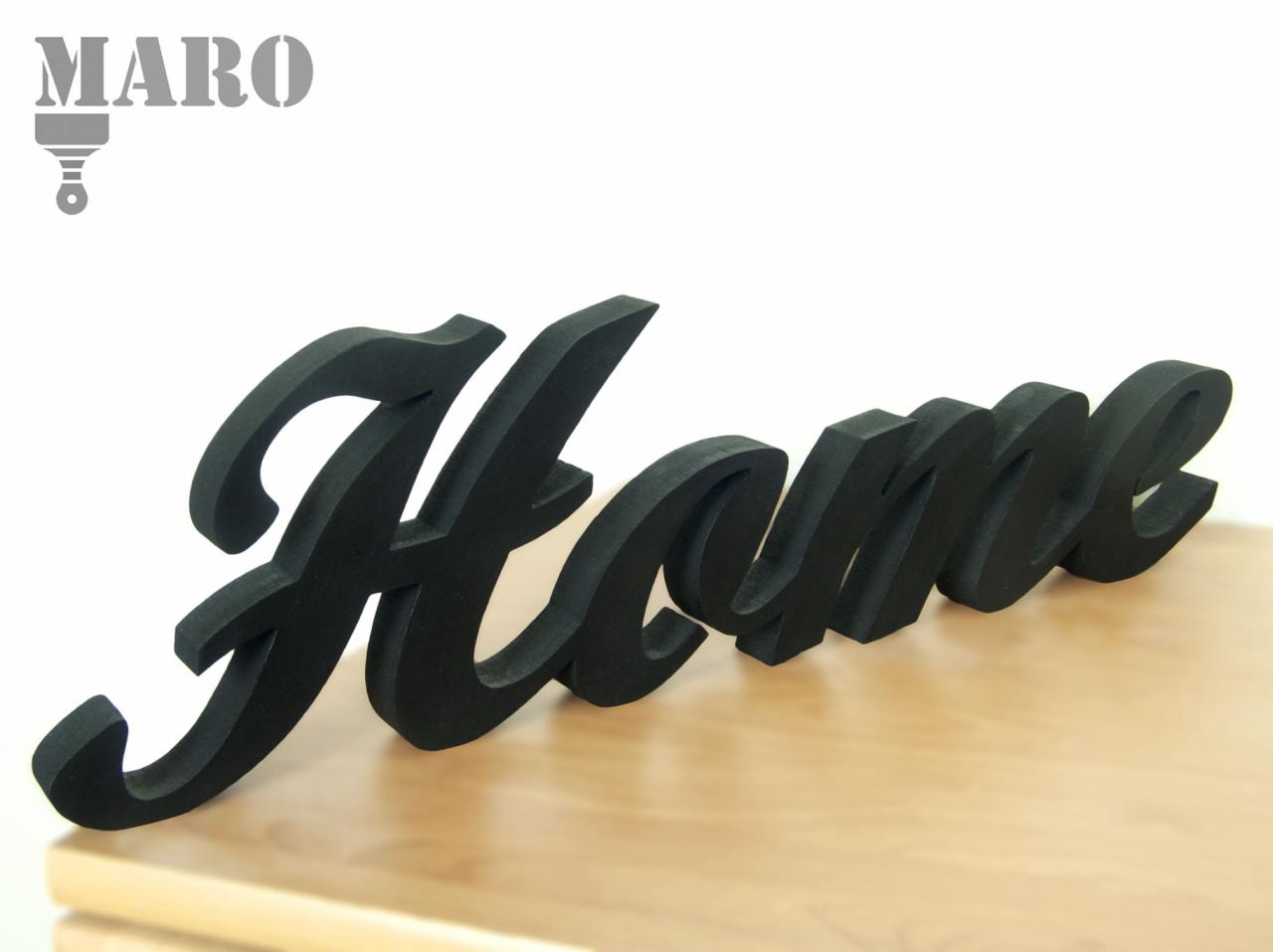 Czarne, trójwymiarowe litery tworzące słowo 'Home' na jasnym drewnianym blacie, z logo firmy MARO w lewym górnym rogu.