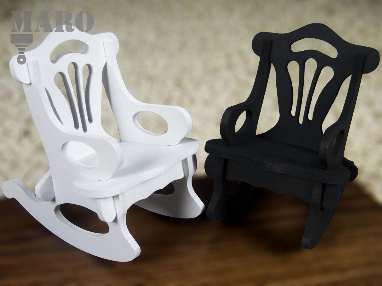 Dwa miniaturowe bujane krzesła, jedno białe, drugie czarne, stojące na drewnianej powierzchni, prezentowane na rozmytym tle.
