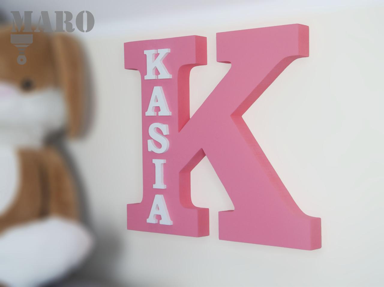 Różowa, drewniana litera K z białym napisem 'KASIA' na białej ścianie, w tle zamazany pluszowy królik.