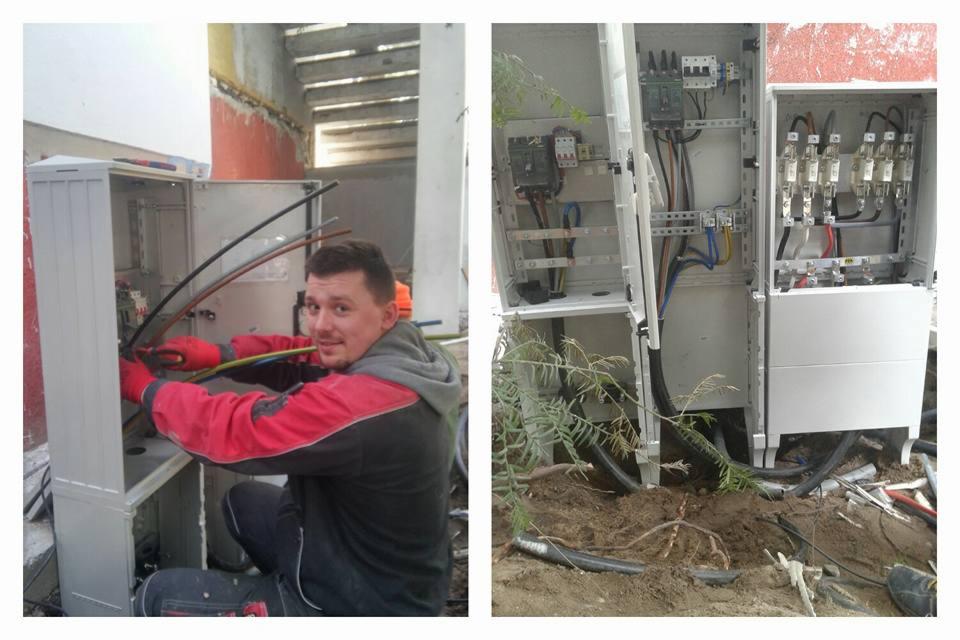 Mężczyzna w czerwonej bluzie i rękawicach łączy przewody w otwartej skrzynce elektrycznej, obok widoczna druga skrzynka z zainstalowanymi elementami, kable wychodzące z ziemi.