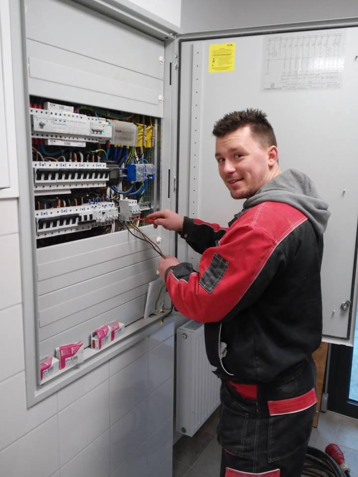 Elektryk w czerwono-czarnym ubraniu roboczym podłącza przewody w szarej skrzynce rozdzielczej w pomieszczeniu wyłożonym białymi płytkami.