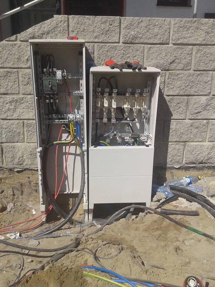 Dwie otwarte skrzynki elektryczne z widocznymi przewodami, bezpiecznikami i elementami montażowymi, umieszczone na ziemi przy ścianie budynku z kamienia, rękawice robocze i śrubokręt na górze...