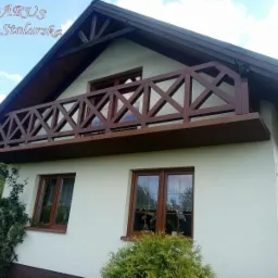 Elewacja domu z drewnianym balkonem o konstrukcji kratownicowej, widoczne brązowe ramy okienne i ciemne orynnowanie.