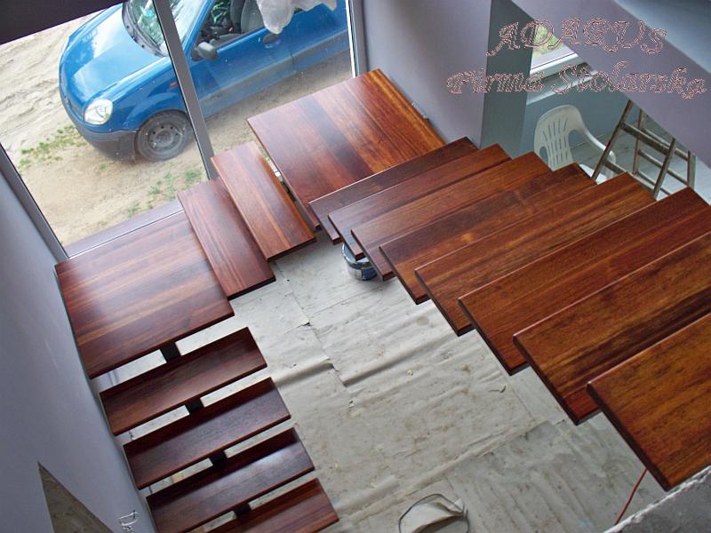 Widok z góry na częściowo zamontowane, lakierowane schody z ciemnego drewna z metalowymi wspornikami, widoczne zabezpieczone podłoże i samochód za oknem.