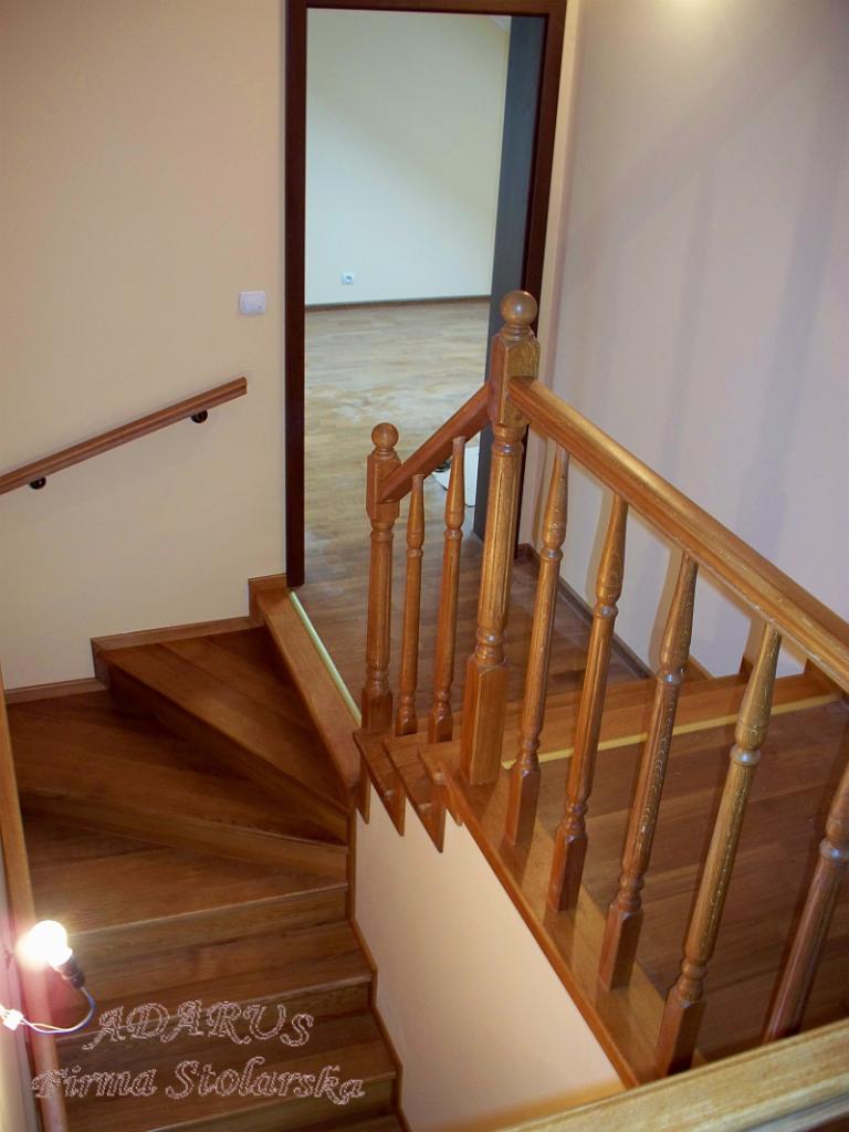 Widok z góry na drewniane schody z balustradą, z widocznym logo firmy stolarskiej na pierwszym stopniu oraz otwartymi drzwiami w tle.