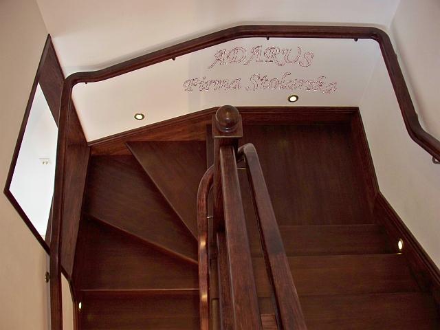Klatka schodowa z ciemnego drewna z oświetleniem punktowym wbudowanym w stopnie, widok od dołu z niskiej perspektywy, na ścianie widoczny napis 'ADARUS Firma Stolarska'.