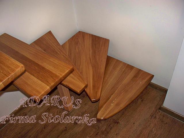 Kątowe ujęcie drewnianych stopni schodowych z widocznym usłojeniem drewna i podpórką, na podłodze z paneli. Na zdjęciu widoczny znak wodny firmy stolarskiej.