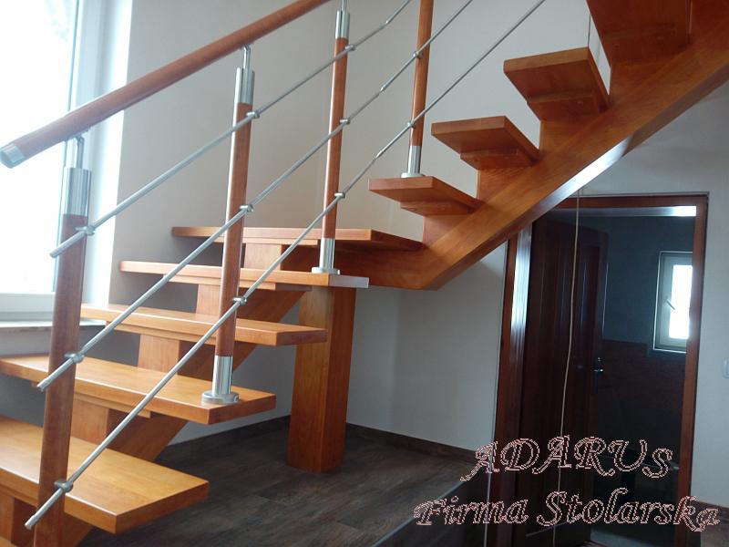 Drewniane schody samonośne z balustradą ze stalowych linek, widoczne od dołu, z ciemną podłogą i drzwiami w tle.