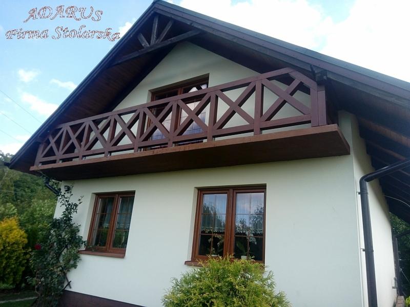 Elewacja domu z drewnianym balkonem o konstrukcji kratownicowej, widoczne brązowe ramy okienne i ciemne orynnowanie.