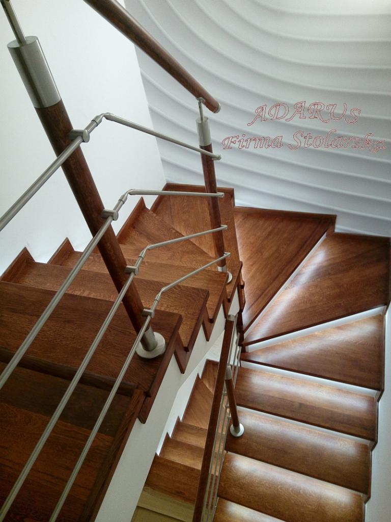 Spiralne schody z ciemnego drewna z metalowymi balustradami, widok z góry, oświetlenie podkreśla strukturę drewna, napis 'ADARUS Firma Stolarska' w prawym górnym rogu.