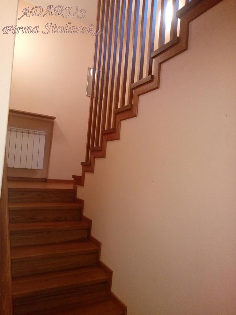 Klatka schodowa z drewnianymi schodami i balustradą z pionowych listew, widoczny grzejnik w tle.