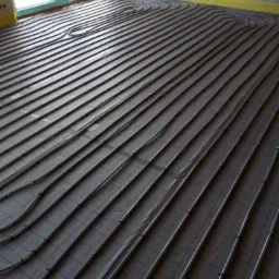 Szare rury ogrzewania podłogowego ułożone równolegle na podłożu z siatką, widoczne żółte taśmy przy ścianach, surowy stan wykończenia wnętrza.