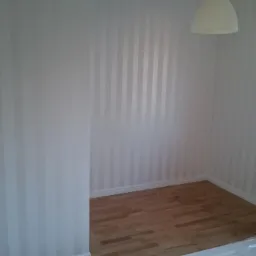 Pokój po remoncie: ściany w pionowe, biało-szare pasy, jasna drewniana podłoga, biały grzejnik pod oknem, widoczna lampa sufitowa i fragment drzwi.