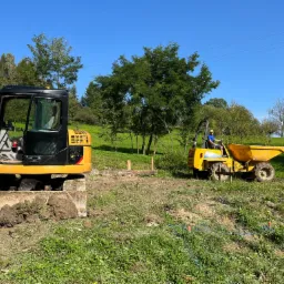 Żółta koparka i wywrotka pracują na zielonym terenie, wykop pod fundamenty w trakcie realizacji, widoczne drewniane znaczniki.