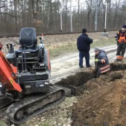 Mała koparka gąsienicowa i grupa robotników w odblaskowych ubraniach pracują przy wykopie w pobliżu torów kolejowych, widoczne świeże ślady pracy w ziemi.