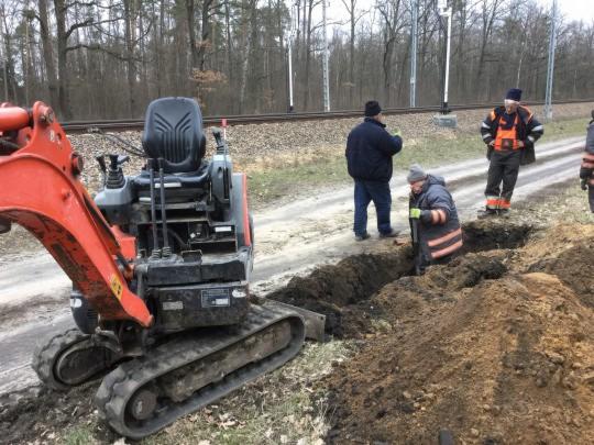 Mała koparka gąsienicowa i grupa robotników w odblaskowych ubraniach pracują przy wykopie w pobliżu torów kolejowych, widoczne świeże ślady pracy w ziemi.