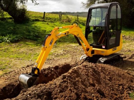Żółta mini koparka JCB w trakcie pracy, tworząca wąski wykop w ziemi na tle zielonego terenu.