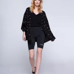 Kobieta o blond włosach pozuje w czarnym topie, spodenkach kolarskich z napisem PILAWSKI i rozpiętej koszuli w małe białe wzory, na nogach czarne sandały na obcasie, na jasnym tle.