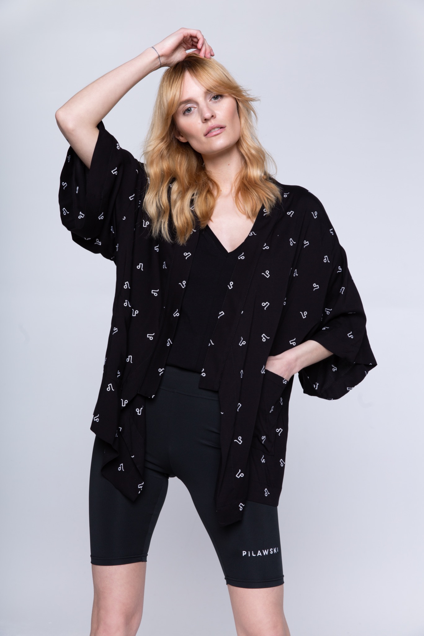 Modelka w czarnym kimonie z białym wzorem zodiakalnym, czarnym topie i legginsach rowerowych z logo 'PILAWSKI' na tle jasnoszarego studia.