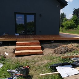 MODEL-JURA - Drewniany taras w trakcie budowy z trzema schodami, widoczna piła do drewna i materiały budowlane na trawie, szara elewacja domu w tle.