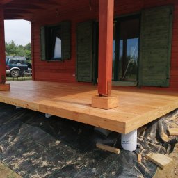 MODEL-JURA - Surowy, drewniany taras w trakcie budowy, wsparty na białych, plastikowych słupach, z widoczną czarną geowłókniną, na tle czerwonego domu z zielonymi okiennicami.