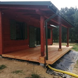 MODEL-JURA - Czerwony drewniany domek z zadaszonym tarasem z jasnego drewna, widoczne słupy podtrzymujące dach i rynna, teren wokół wyłożony czarną folią, w tle drzewa.