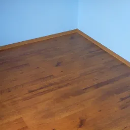 Ułożone panele podłogowe w odcieniach brązu, widoczne łączenia i listwa przypodłogowa przy jasnoniebieskiej ścianie w rogu pomieszczenia.