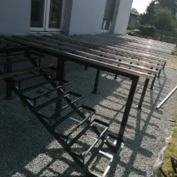 Metalowa konstrukcja pod taras z przygotowanymi schodami, widoczne spawane elementy i podłoże z kamyków.