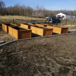 Drewniane skrzynie ogrodowe ustawione na ziemi, wyłożone czarną folią, w tle dom jednorodzinny i samochód terenowy.