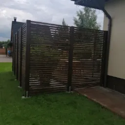 Nowoczesna, pozioma, brązowa przesłona z listew, oddzielająca przestrzeń, zamontowana na metalowych wspornikach, widok z zewnątrz na tle trawnika i budynku z rynną.