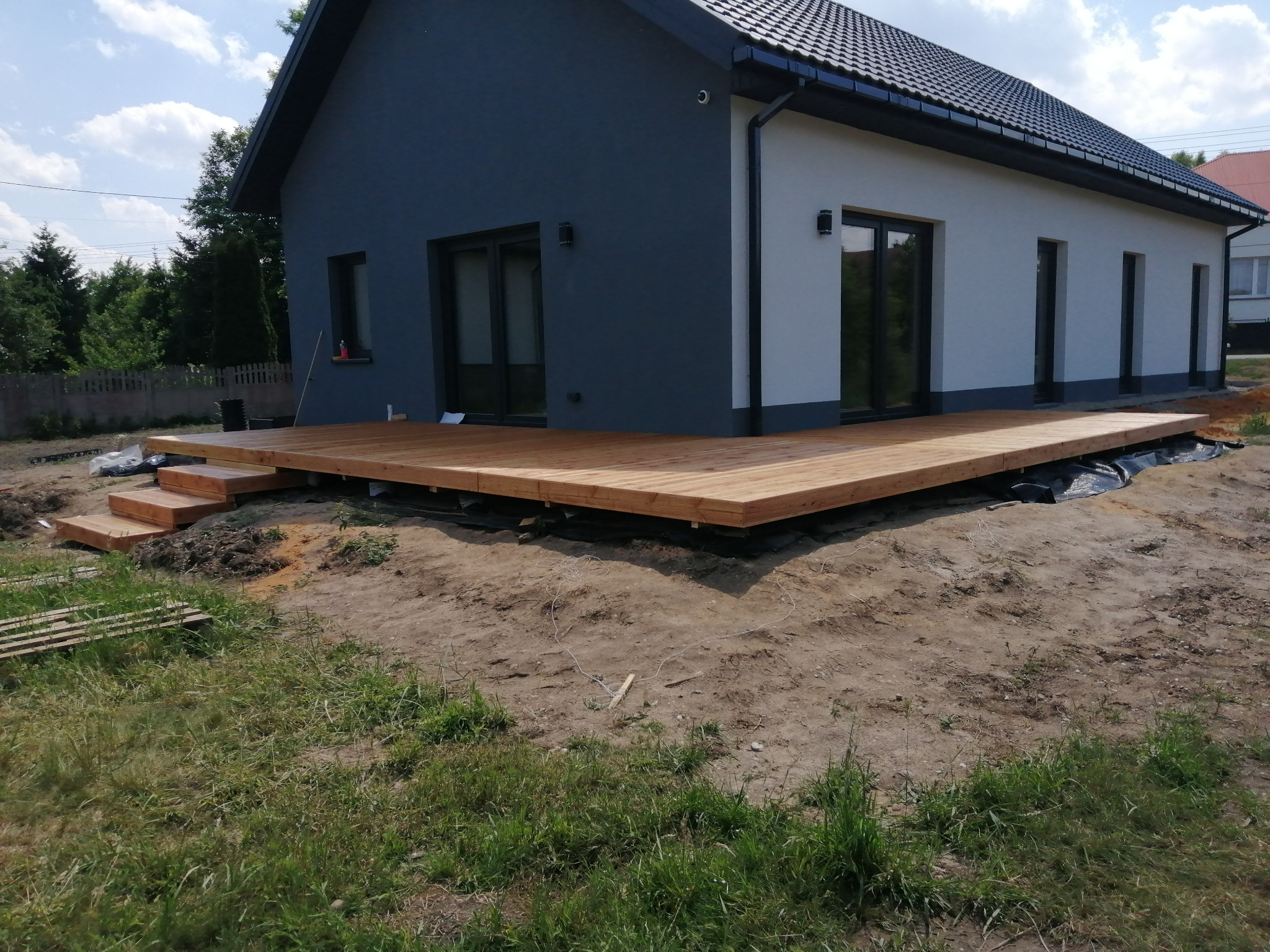 Nowoczesny dom z szarą elewacją i drewnianym tarasem w trakcie budowy, widoczne deski i czarna folia izolacyjna, ziemia wokół i trawa.