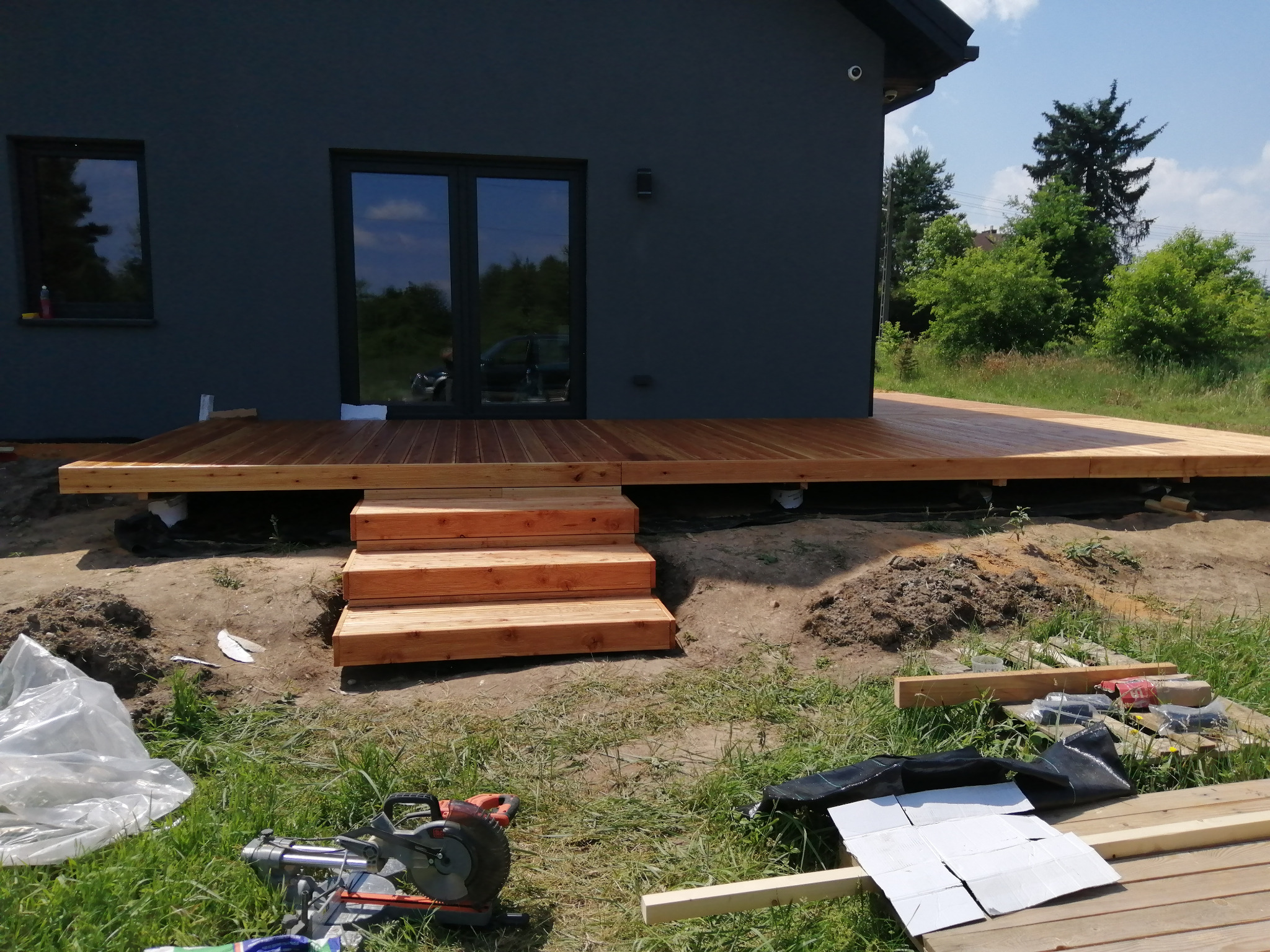 Drewniany taras w trakcie budowy z trzema schodami, widoczna piła do drewna i materiały budowlane na trawie, szara elewacja domu w tle.
