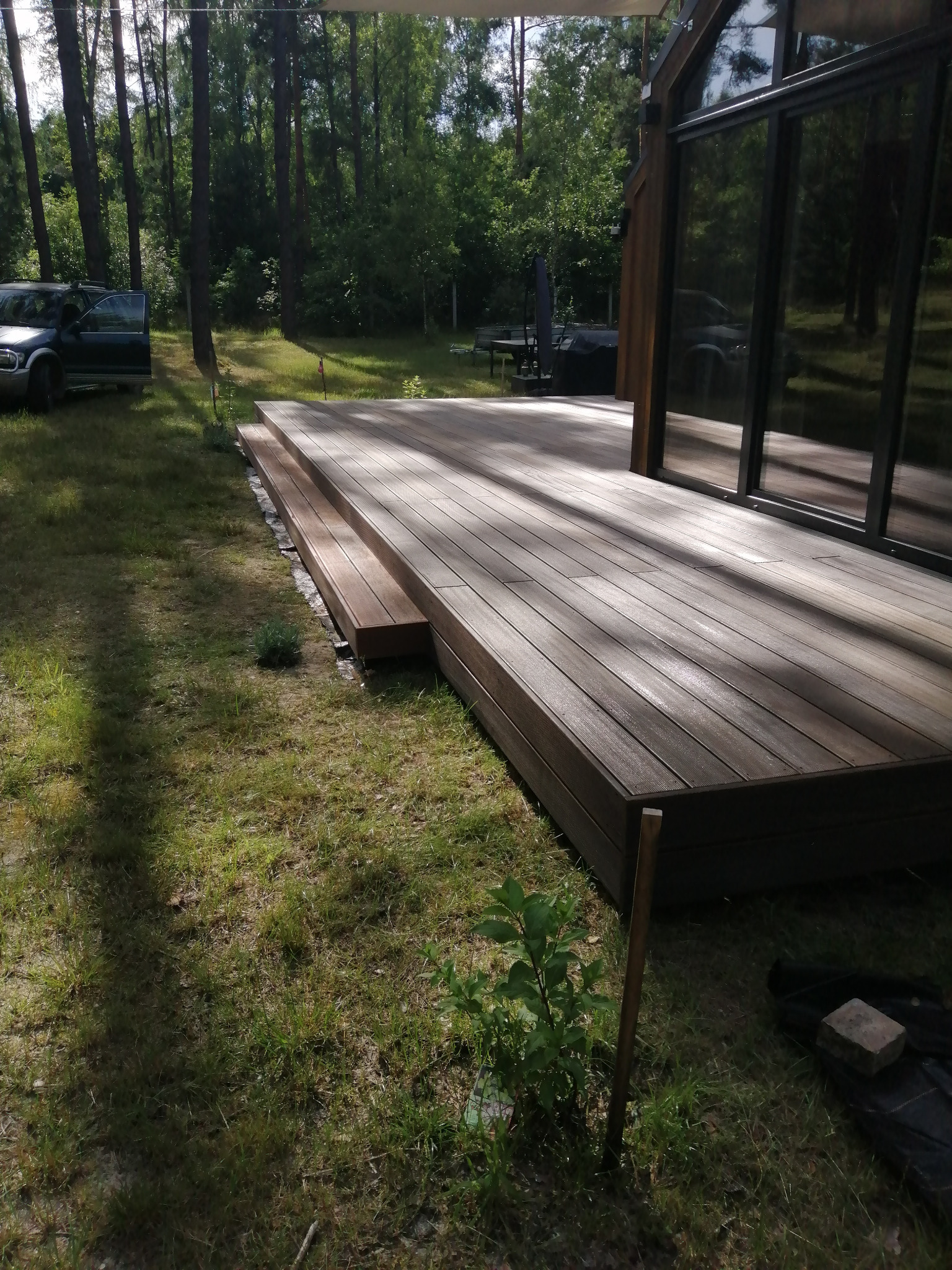 Nowoczesny taras kompozytowy z dwoma stopniami, przylegający do budynku z dużymi oknami, otoczony zielonym trawnikiem i lasem.