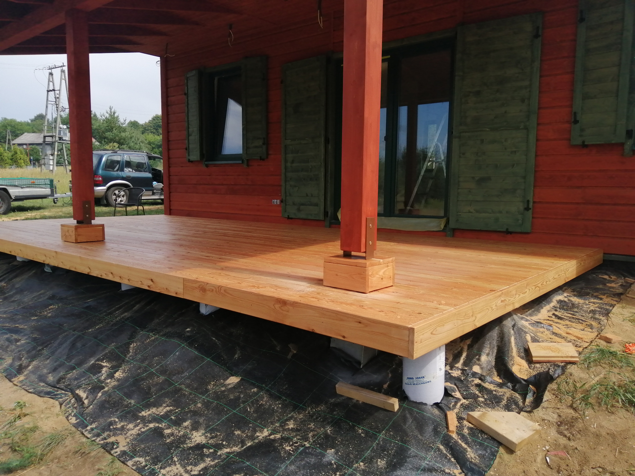 Surowy, drewniany taras w trakcie budowy, wsparty na białych, plastikowych słupach, z widoczną czarną geowłókniną, na tle czerwonego domu z zielonymi okiennicami.