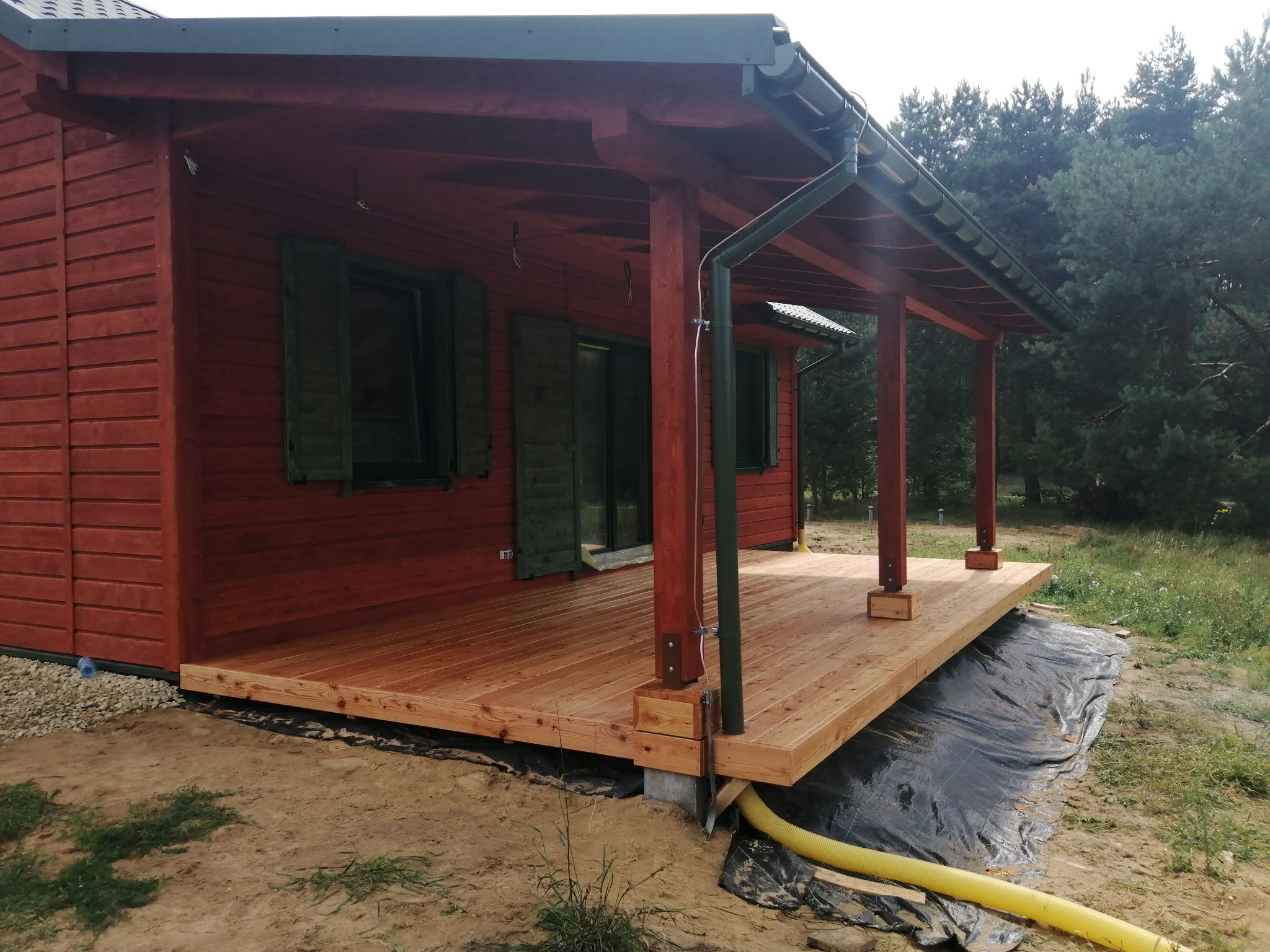 Czerwony drewniany domek z zadaszonym tarasem z jasnego drewna, widoczne słupy podtrzymujące dach i rynna, teren wokół wyłożony czarną folią, w tle drzewa.