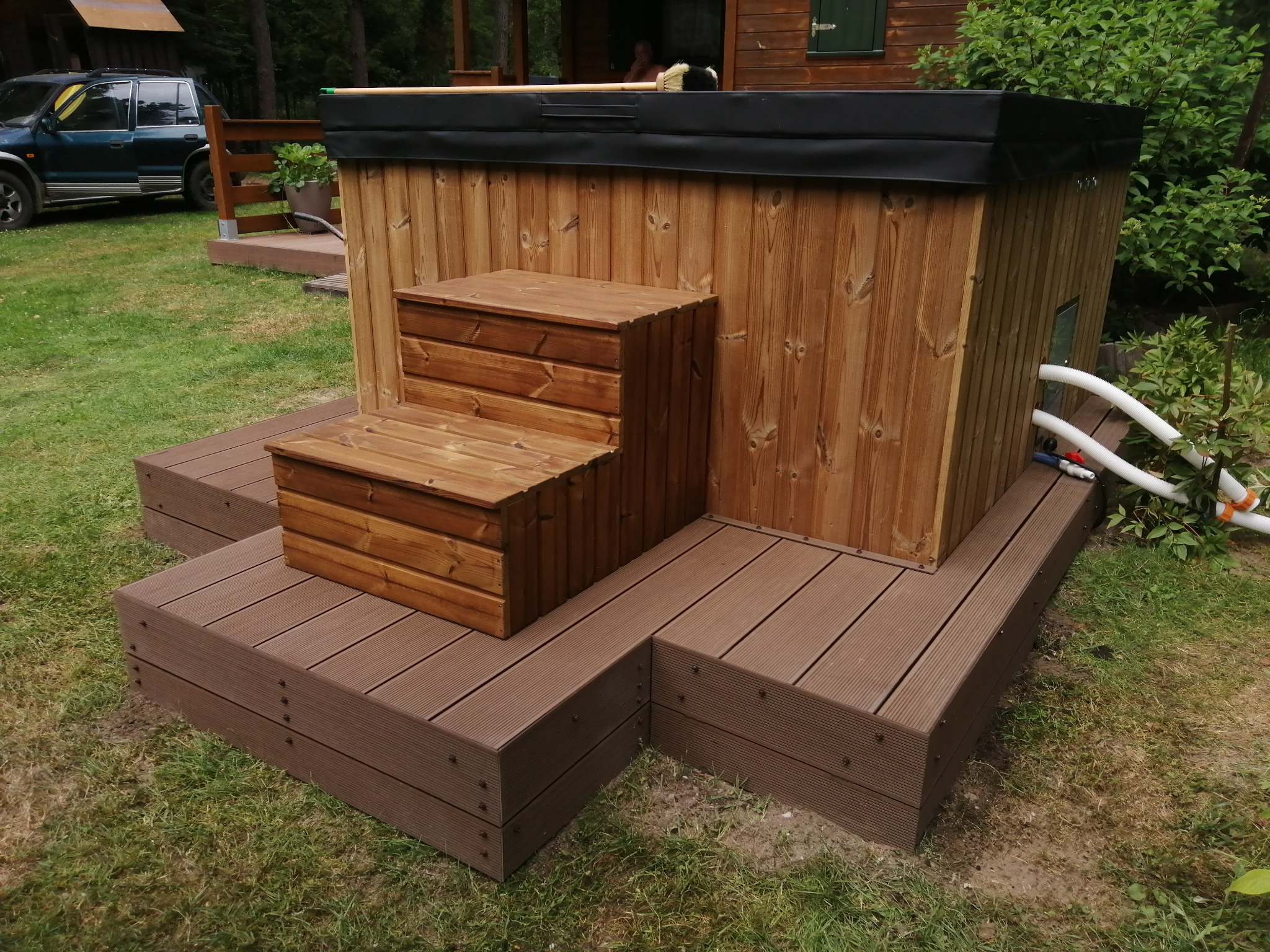 Drewniana wanna z hydromasażem otoczona brązowym tarasem kompozytowym i schodkami, z widocznymi białymi rurami i zielenią w tle.