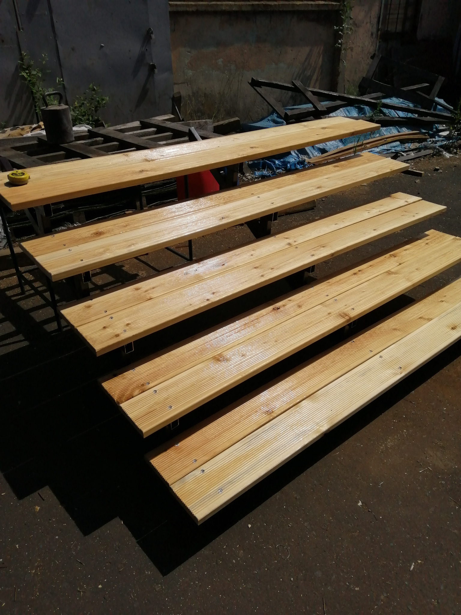 Surowe, drewniane stopnie schodów z widocznymi śrubami, ustawione na metalowej konstrukcji w warsztatowym otoczeniu, obok leży miarka.