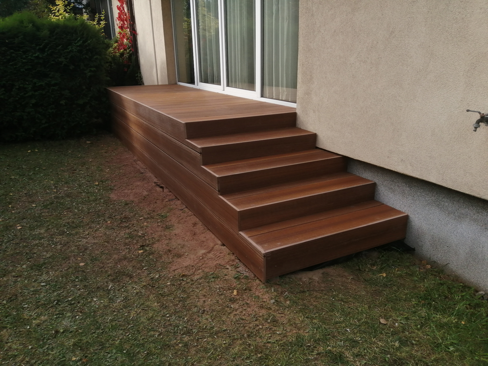 Brązowy taras kompozytowy z czterema stopniami prowadzący do przeszklonych drzwi balkonowych, widok z poziomu trawnika.