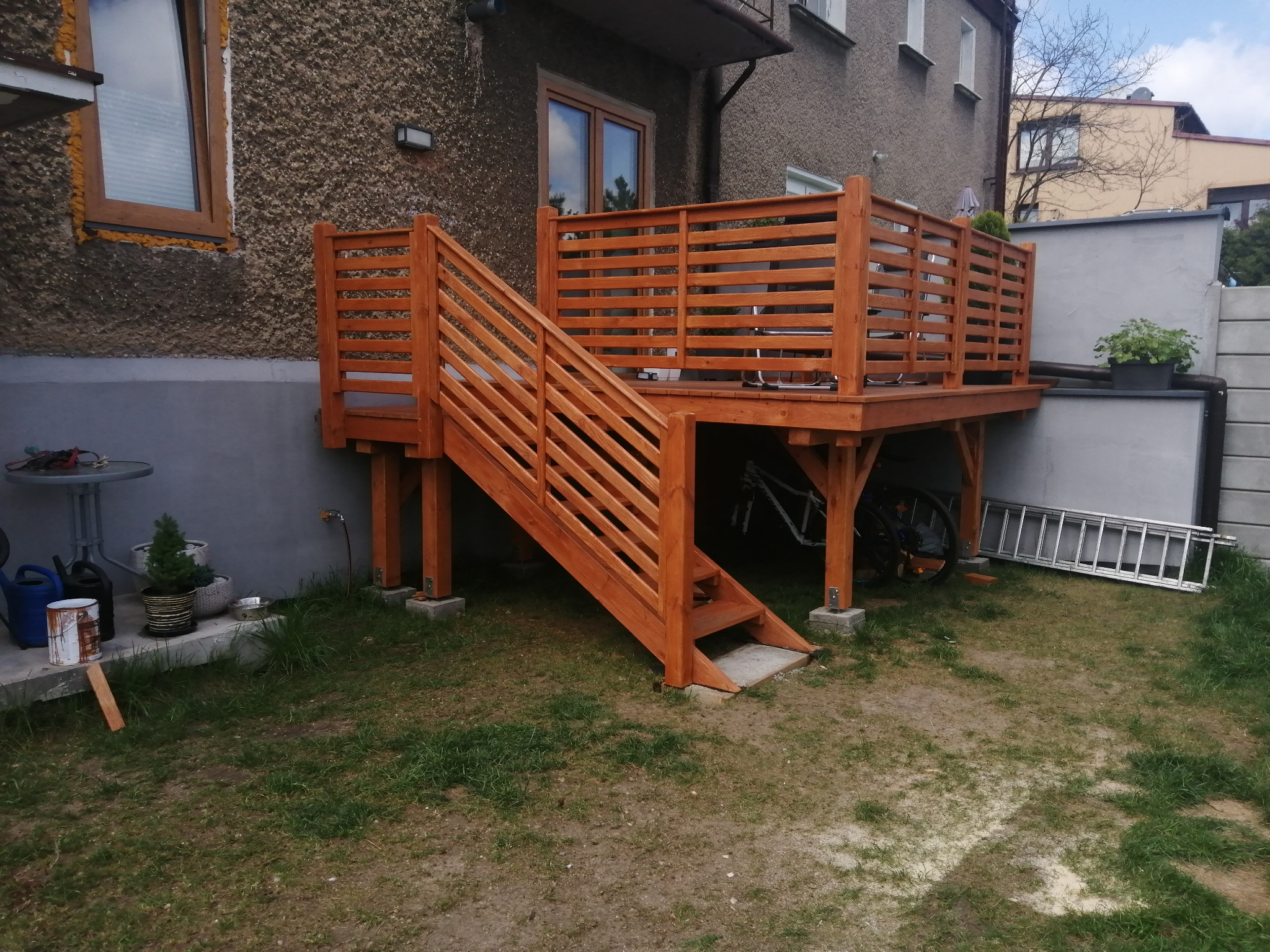 Drewniany taras z balustradą i schodami przy budynku mieszkalnym. Konstrukcja na podwyższeniu, z widocznymi podporami. Na tarasie doniczki z roślinami.